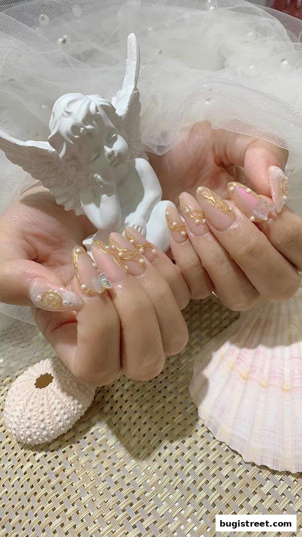 Close-up of hands with intricate pearl and gold nail art, styled with a cherub statue and seashells.