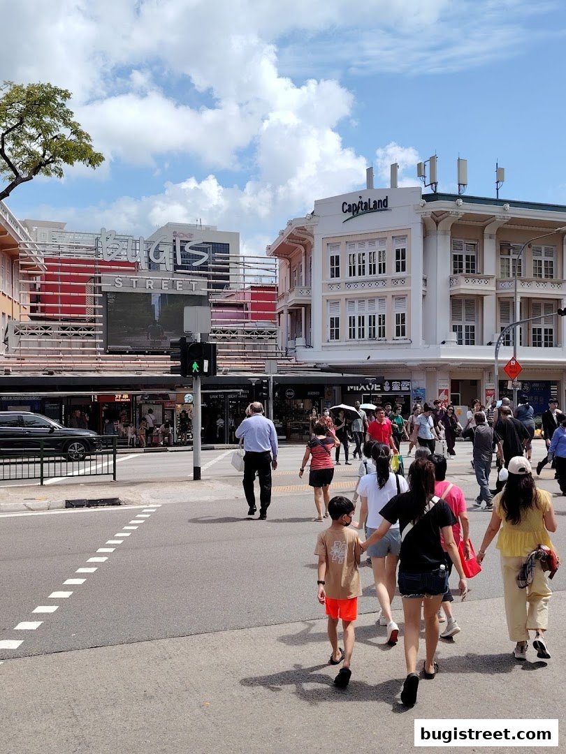 People crossing a street in front of shops and buildings on a sunny day.