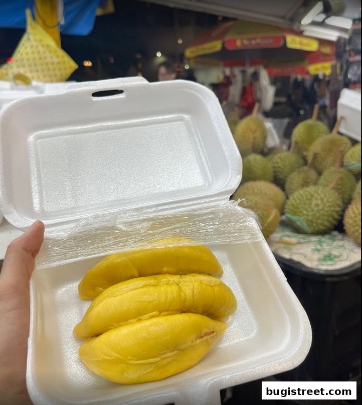 A hand holds a takeaway container filled with fresh, ripe durian fruit.