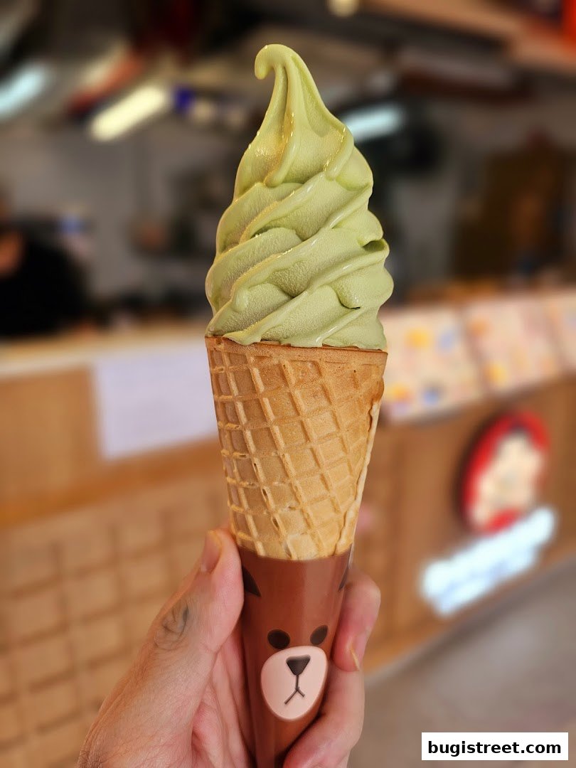 A hand holds a green soft-serve ice cream cone with a cute bear-themed wrapper.