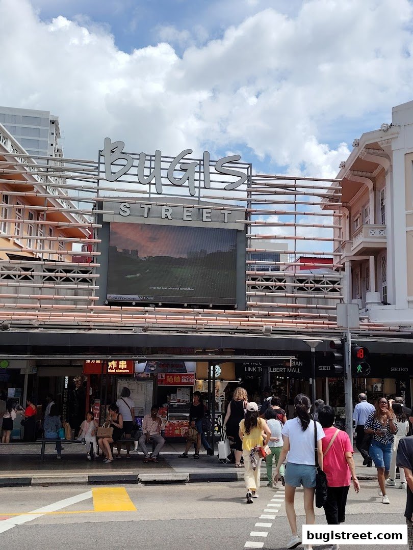 People walking on a street in front of Bugis Street with shops and a large screen.