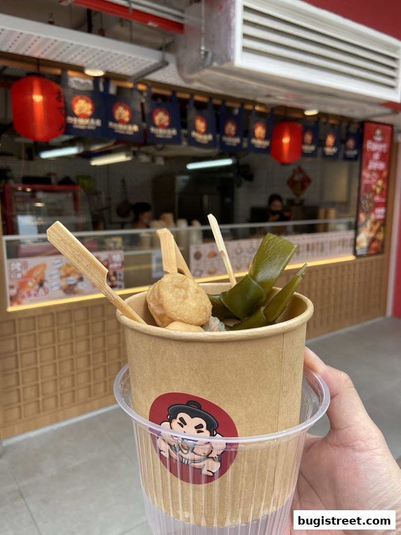 A hand holds a cup of Oden with various ingredients, in front of a shop.