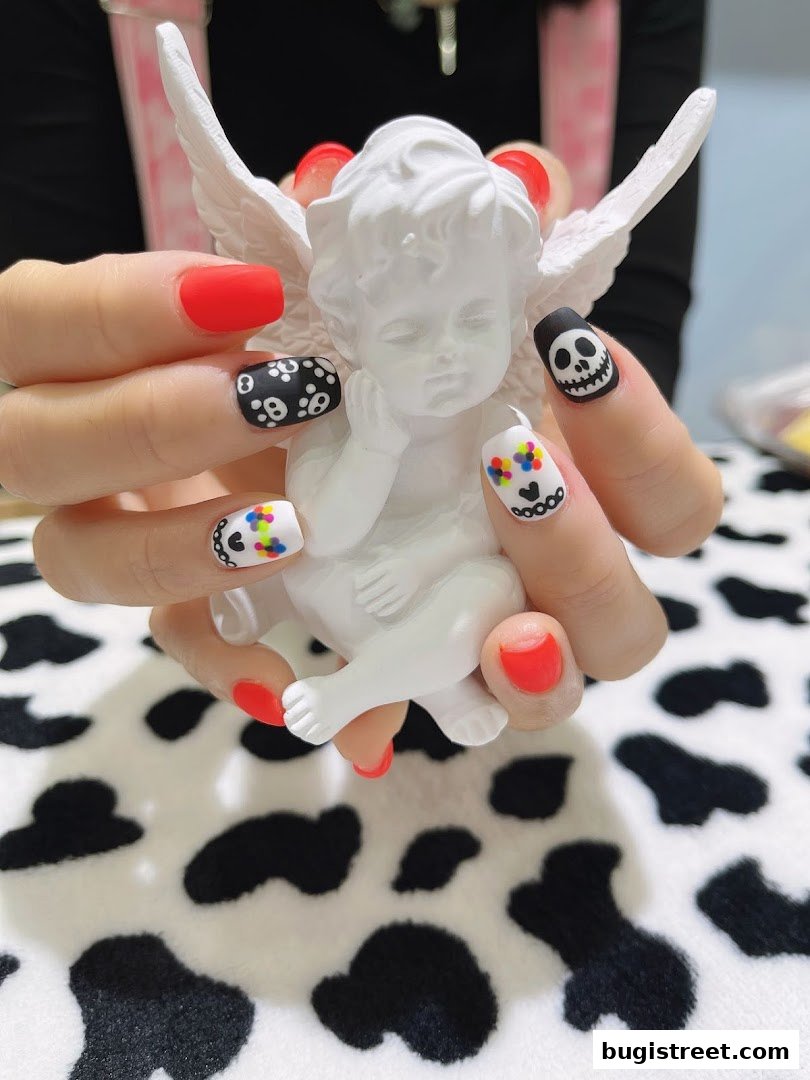 Hands with creative nail art featuring skull designs and bright colors, holding a white cherub statue.