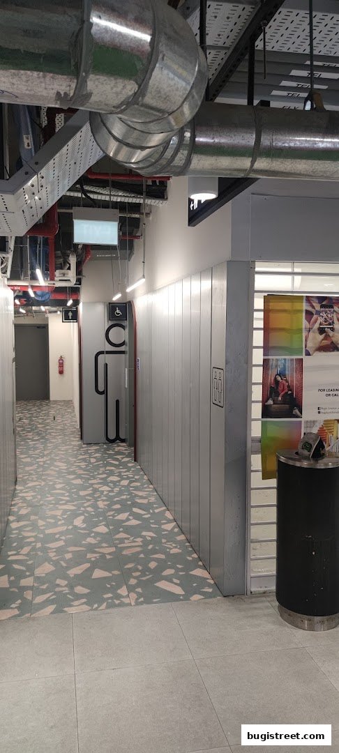 A hallway with exposed pipes, signage for restrooms, and a trash can.