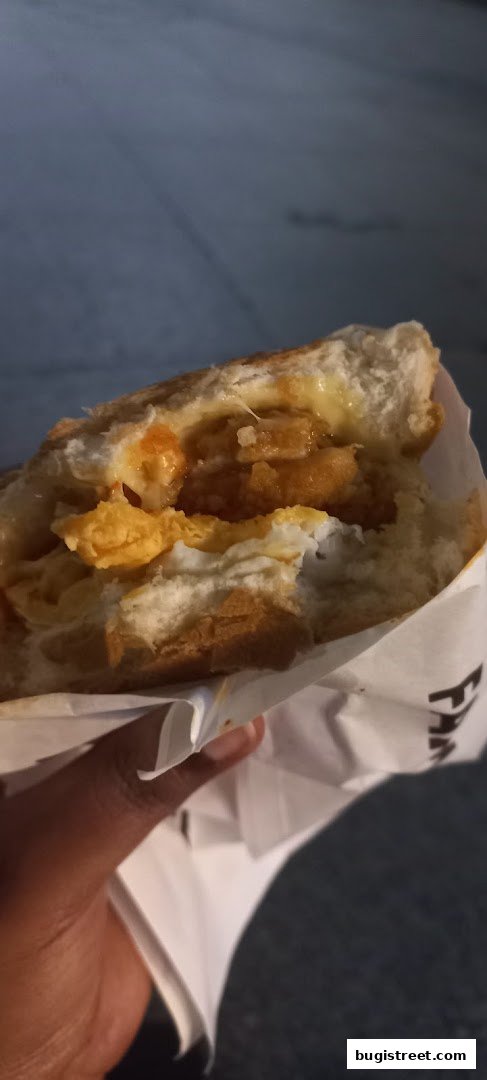 A hand holds a delicious-looking sandwich with fried chicken, egg, and sauce.