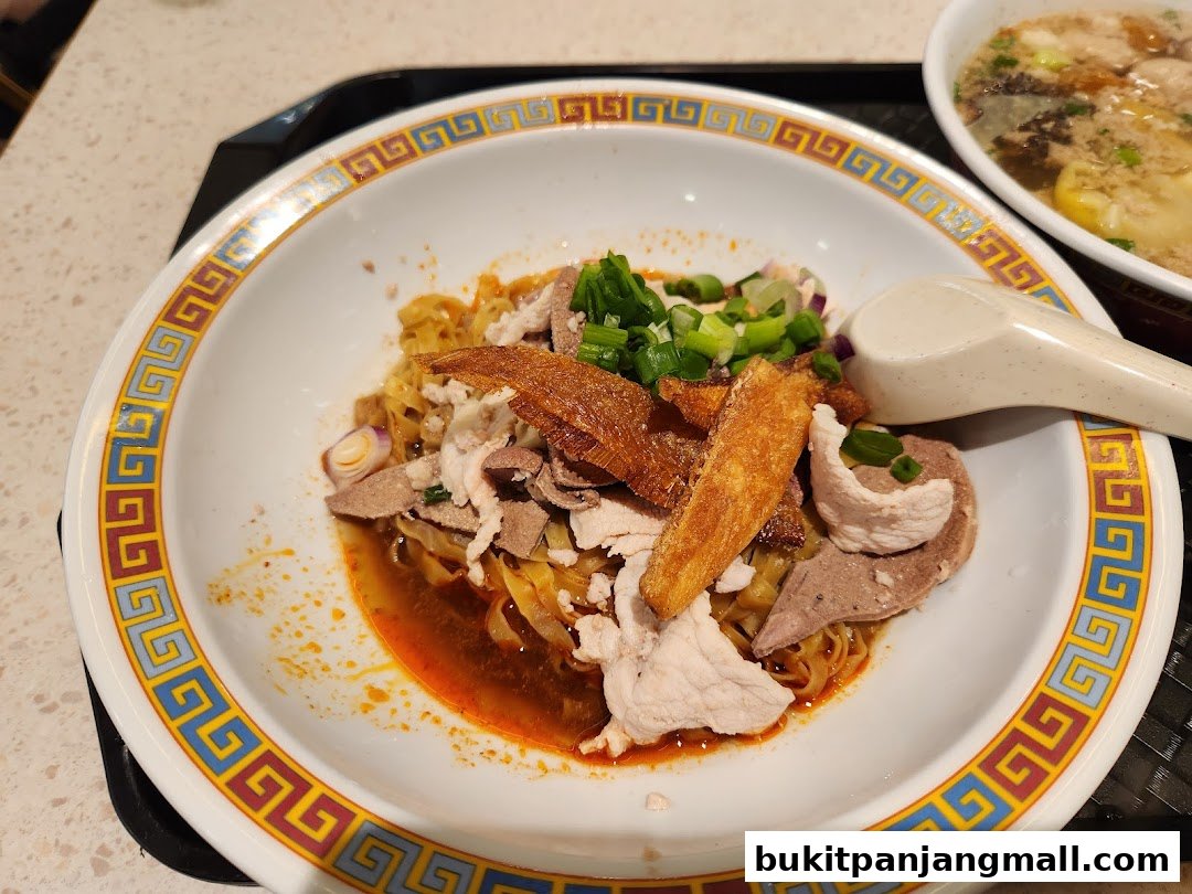 A bowl of noodles with meat and crispy toppings, served with a side of soup.