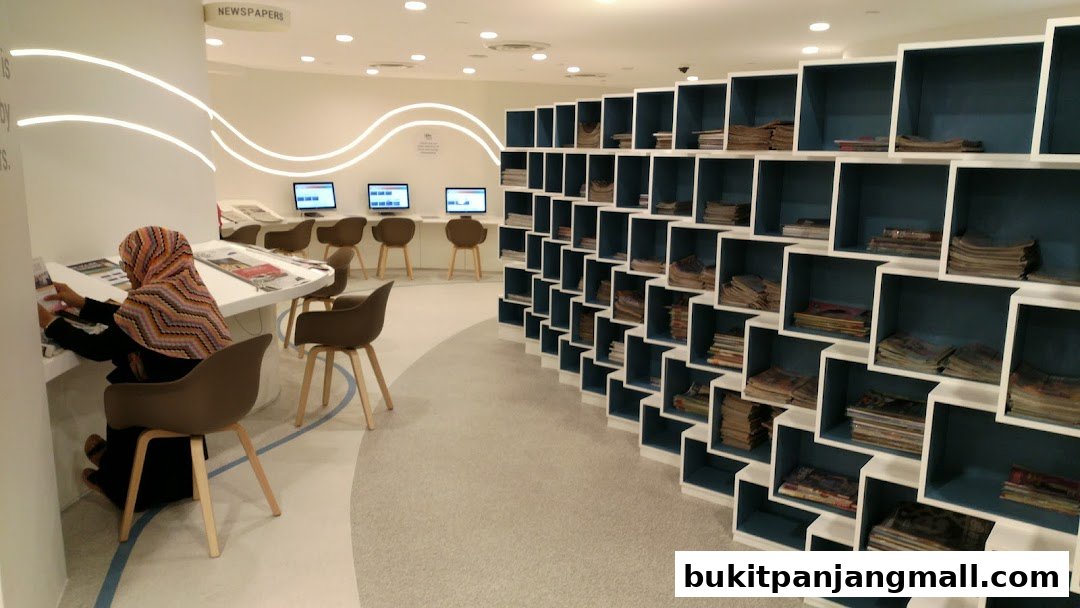 A person reads at a table with computers in the background and a wall of shelves filled with magazines.