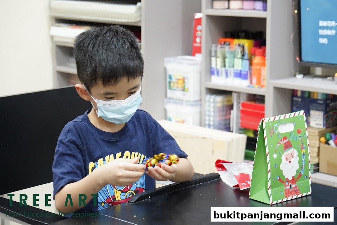 A young boy wearing a mask works on a craft project at a table with art supplies in the background.