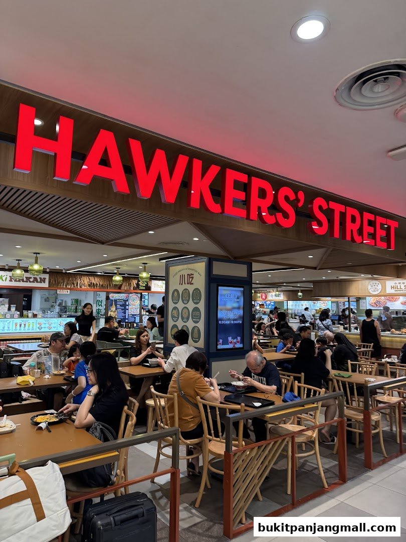 A busy hawker street food court with many diners eating at tables under a large red sign.