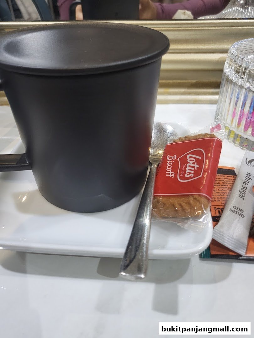 A black mug with a lid sits on a white tray with a spoon and biscuit.