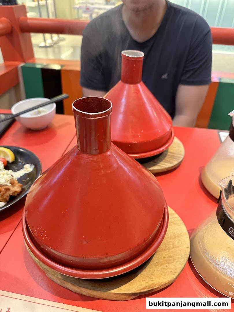 Two steaming red conical pots sit on a red table, with a person in the background.