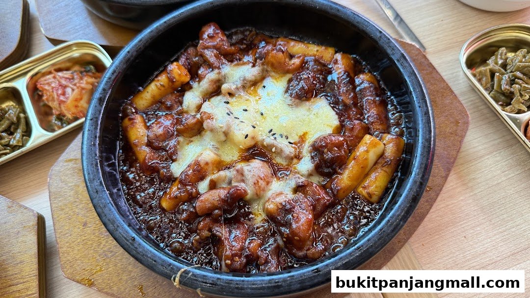 A close-up of a Korean dish with chicken, rice cakes, and melted cheese, served with kimchi and other side dishes.