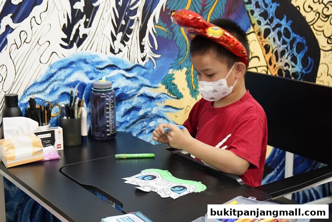 A young boy wearing a snake headband and mask is coloring a drawing at a desk.