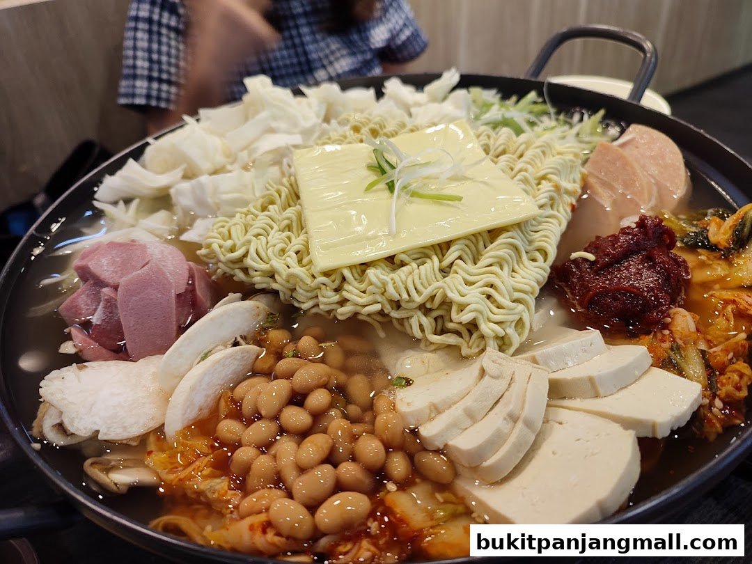 A close-up of a bubbling Korean stew with noodles, cheese, beans, tofu, and ham.