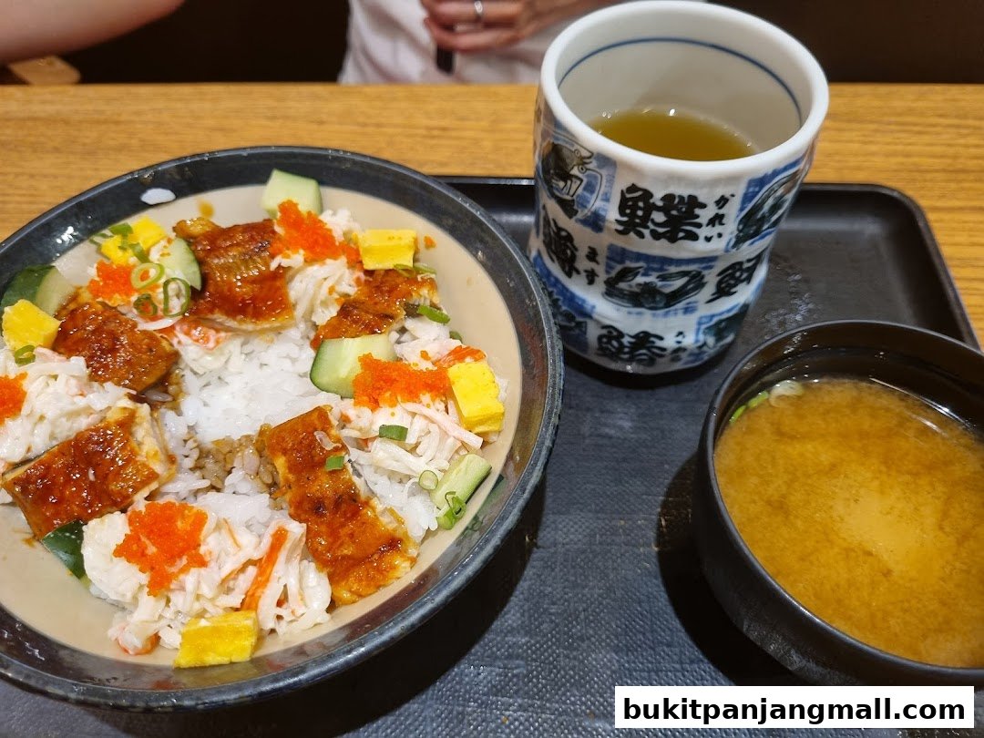 A bowl of Japanese rice dish with grilled eel, crab sticks, cucumber, and roe, served with miso soup and tea.