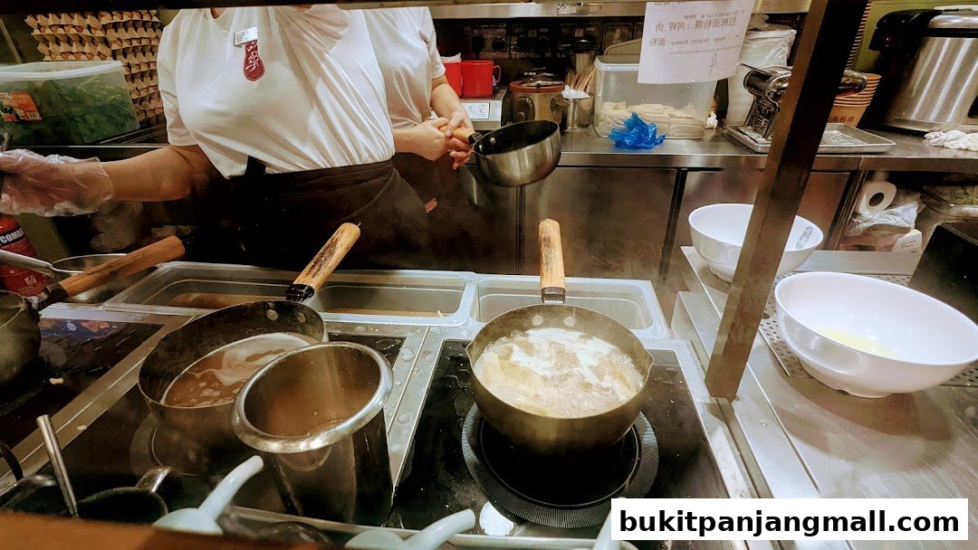 A chef prepares Mee Hoon Kway in a busy kitchen with pots of broth and ingredients.
