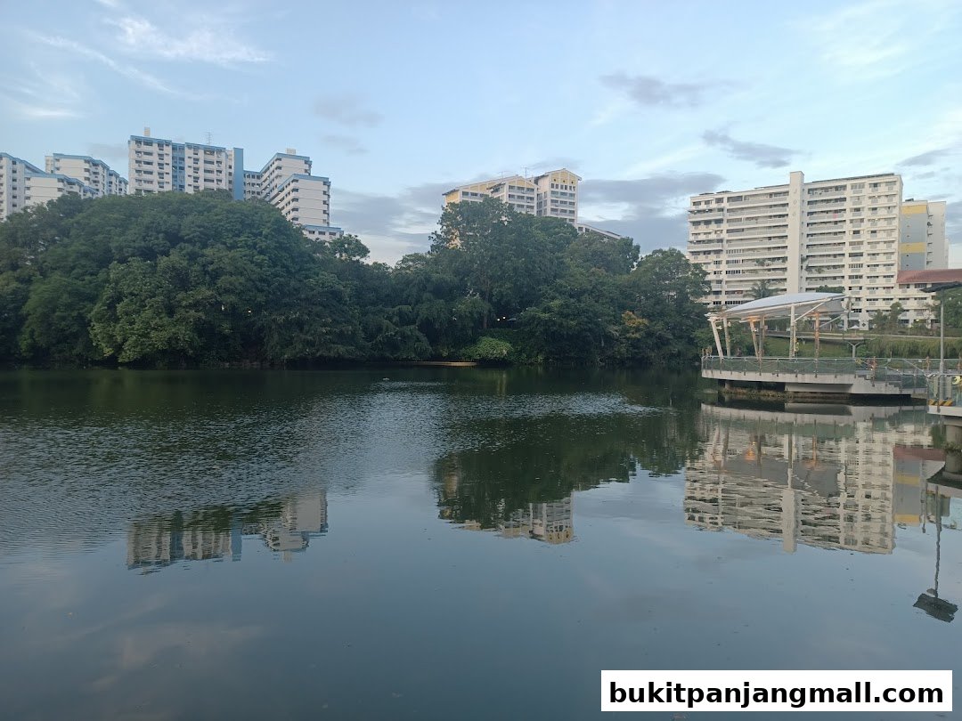 A serene lake with reflections of apartment buildings and lush trees under a cloudy sky.