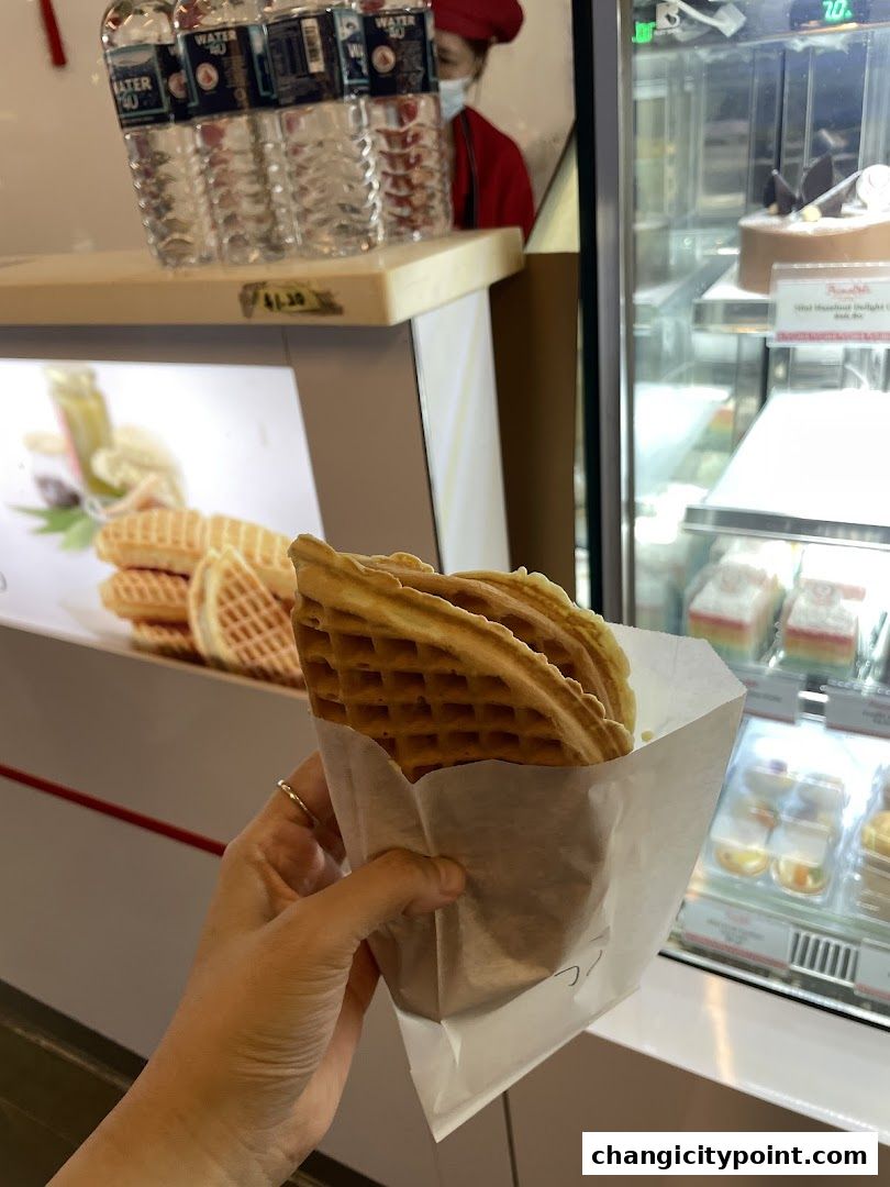 A hand holds a freshly made waffle cone from PrimaDéli, with cakes visible in the display fridge.