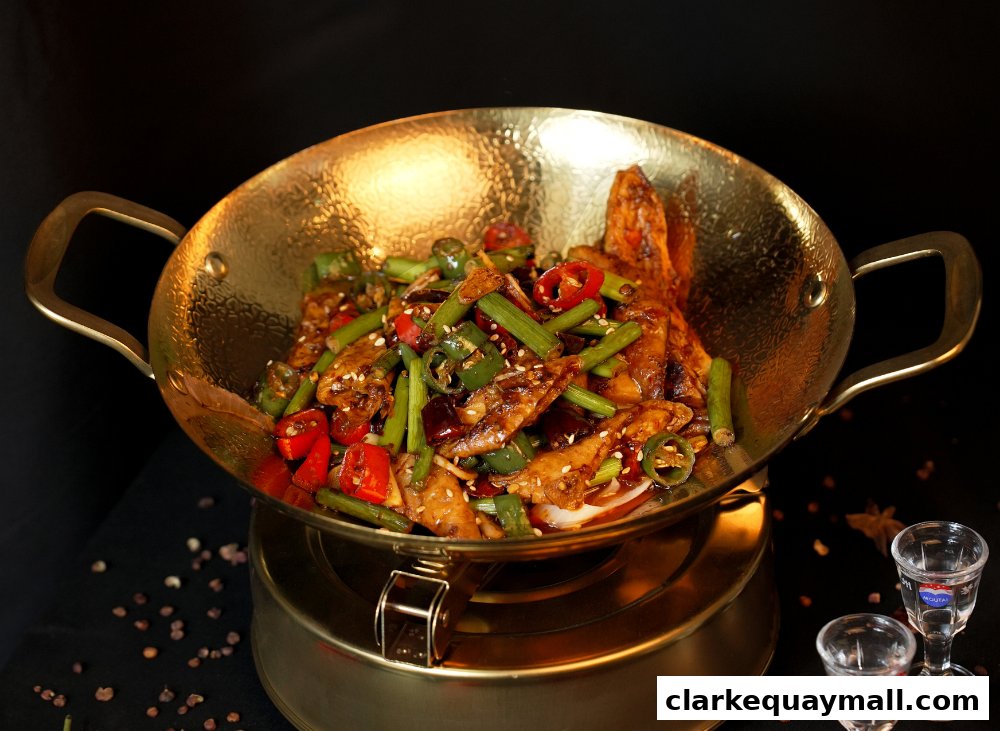 A golden wok filled with spicy chicken wings, chili peppers, and green onions, served with two shot glasses.