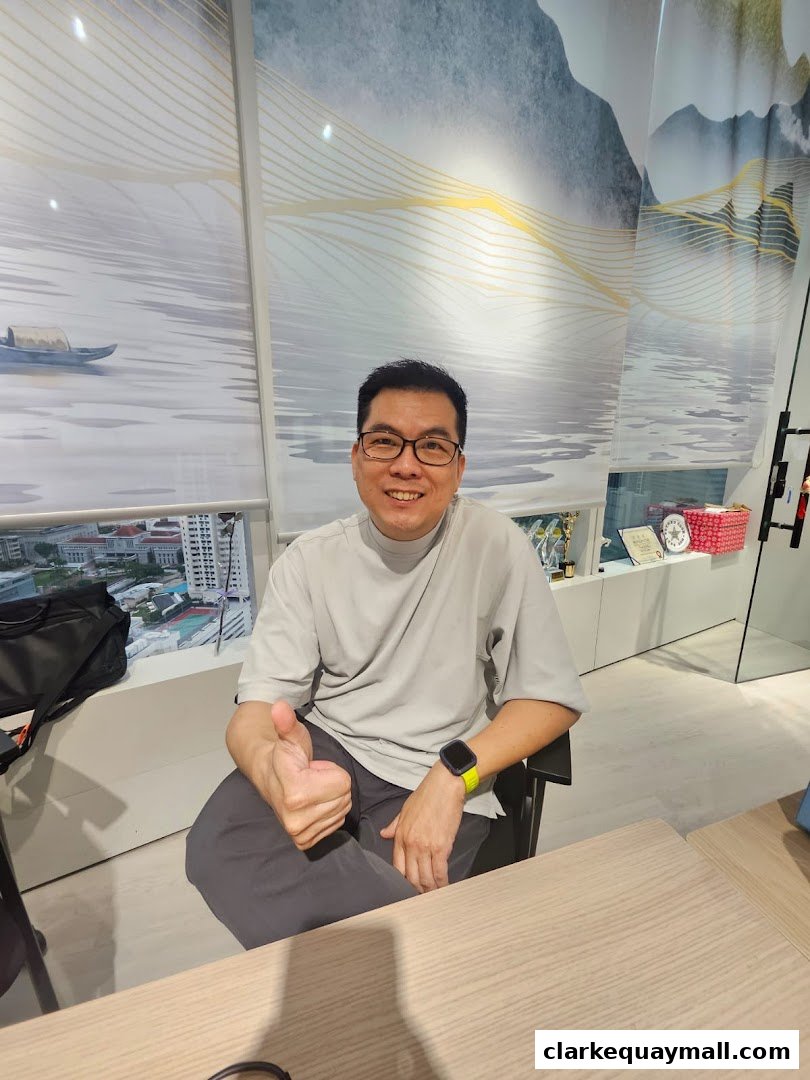 A man in a grey t-shirt gives a thumbs up while sitting at a desk with a scenic backdrop.