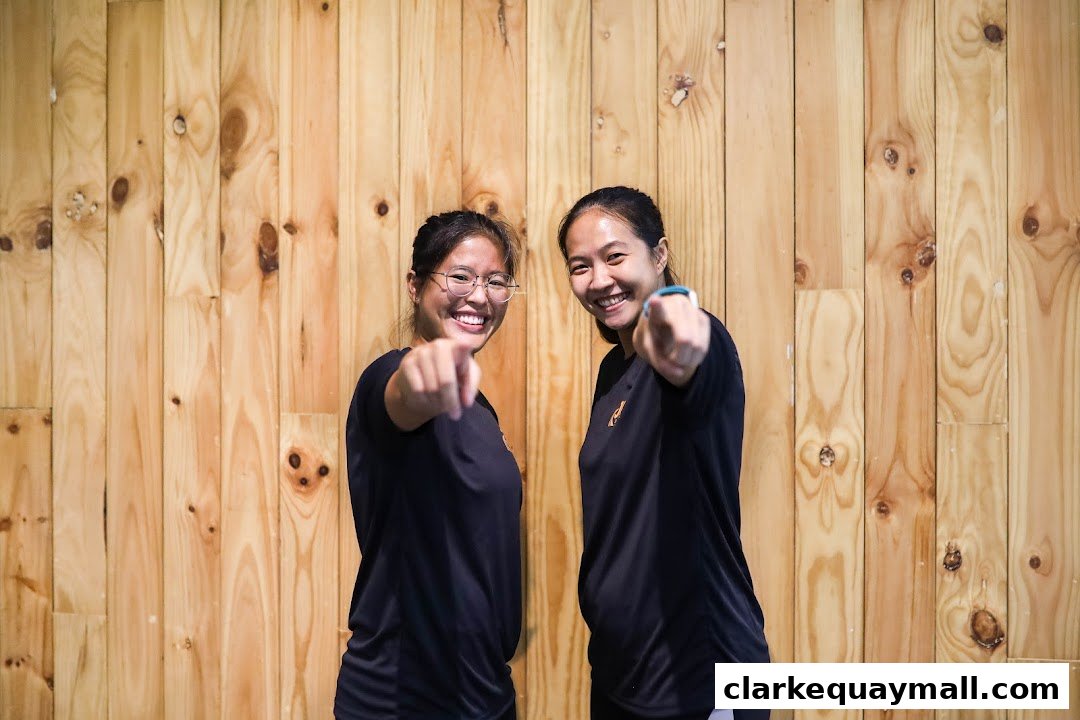 Two smiling women in black t-shirts point forward against a wooden background.