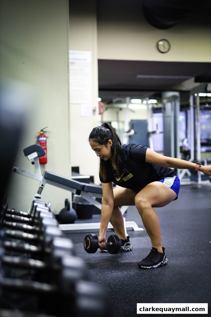 A woman is performing a dumbbell deadlift in a gym, with weights and equipment in the background.