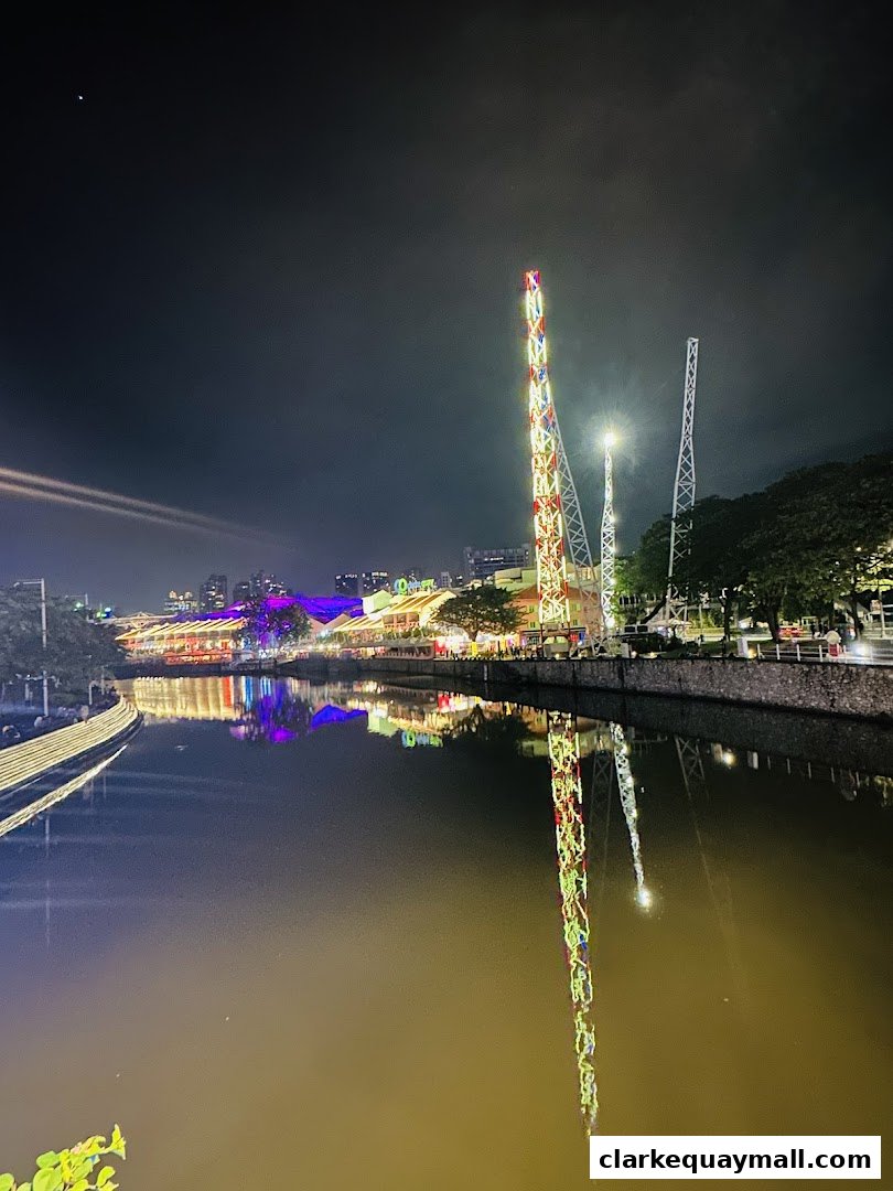 Night view of a river with illuminated buildings and tall amusement rides reflected in the water.