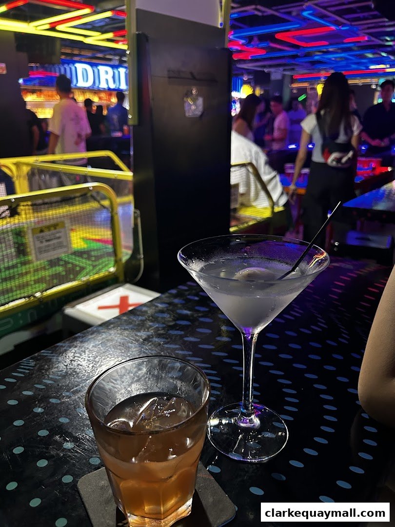 Two cocktails on a table in a lively arcade bar with neon lights and people in the background.
