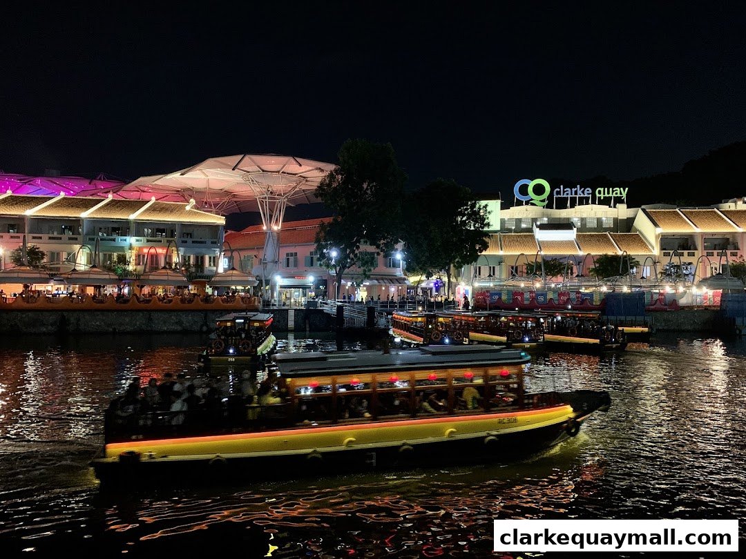 Night view of boats on a river with illuminated buildings and a 'Clarke Quay' sign.