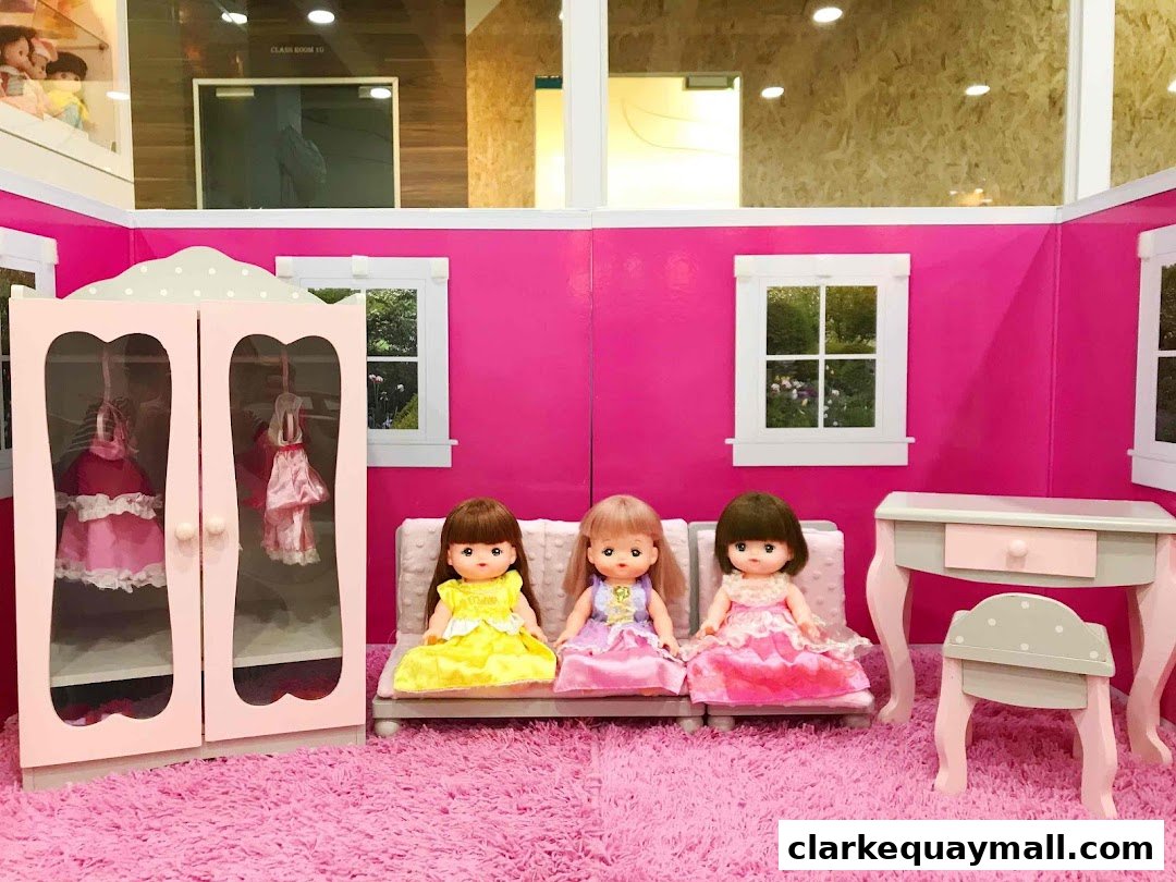 Three dolls are sitting on a pink couch in a room with a doll wardrobe and vanity.