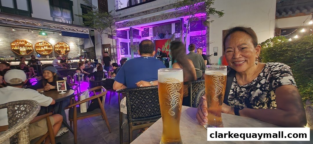 A woman smiles at the camera with two beers in front of her at a lively bar with live music.