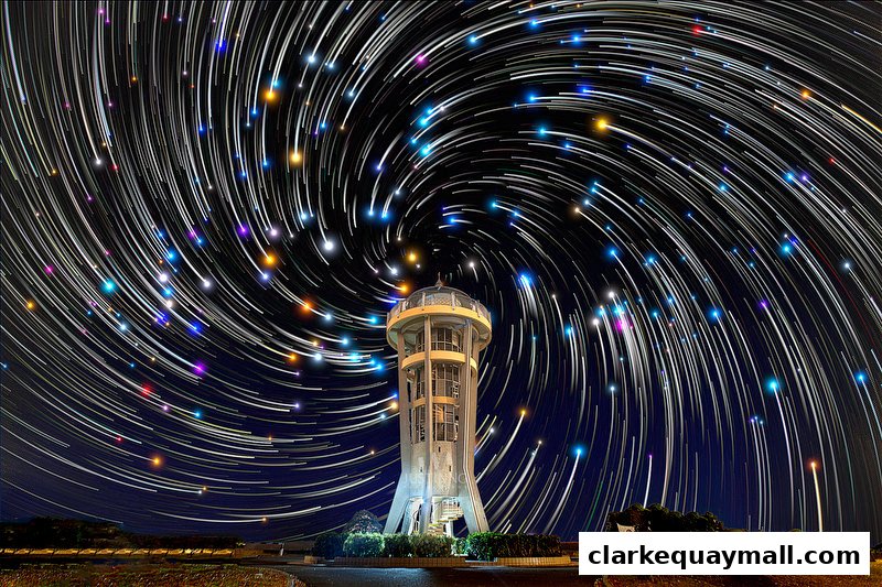 A tower stands under a swirling vortex of star trails at night.