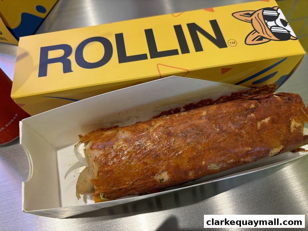 A golden-brown cheese-crusted hot dog in a white paper tray with the 'Rollin Cheese Dogs' logo box behind it.