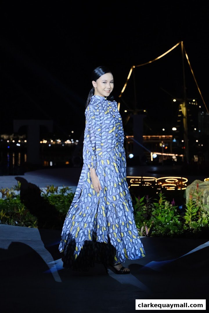 A woman in a patterned dress walks on a stage at night with city lights in the background.