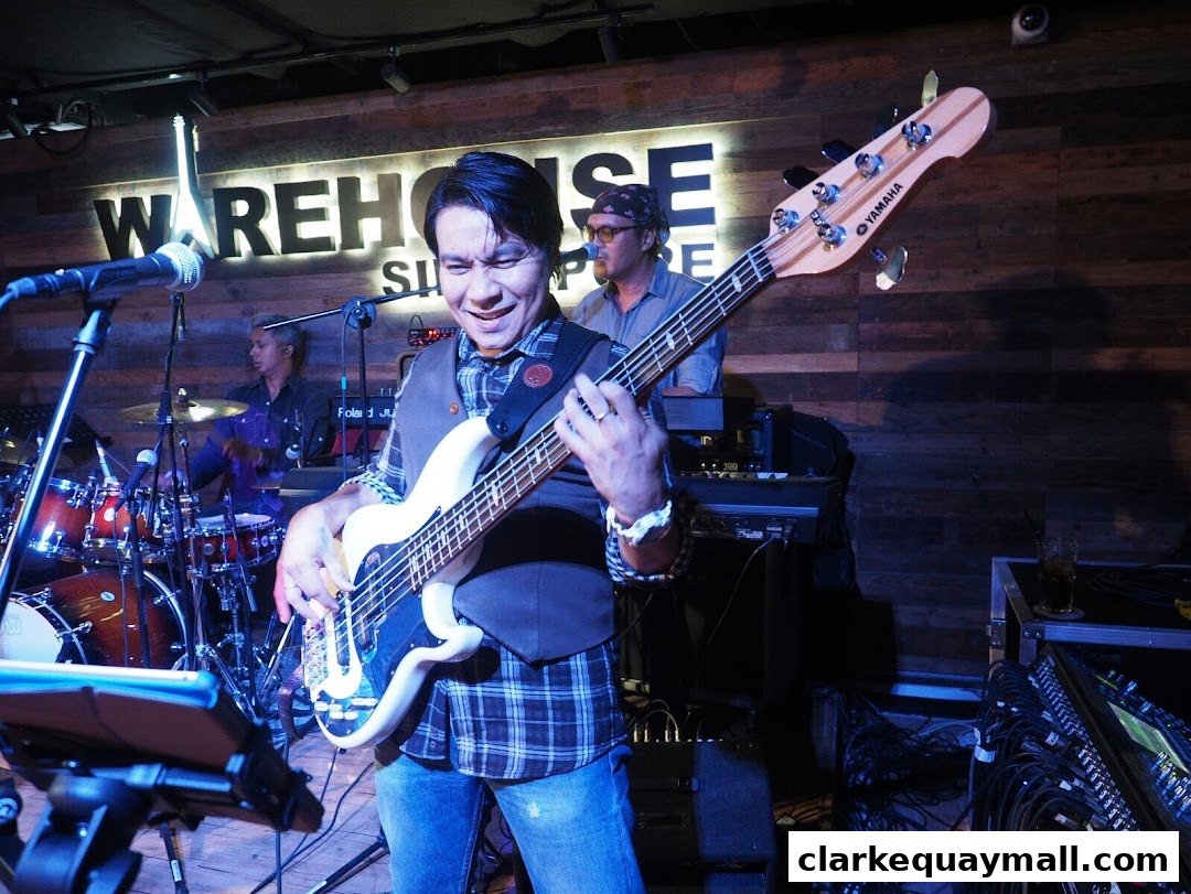 A band performs live music on stage at Warehouse Bar, Singapore, with a bassist in the foreground.