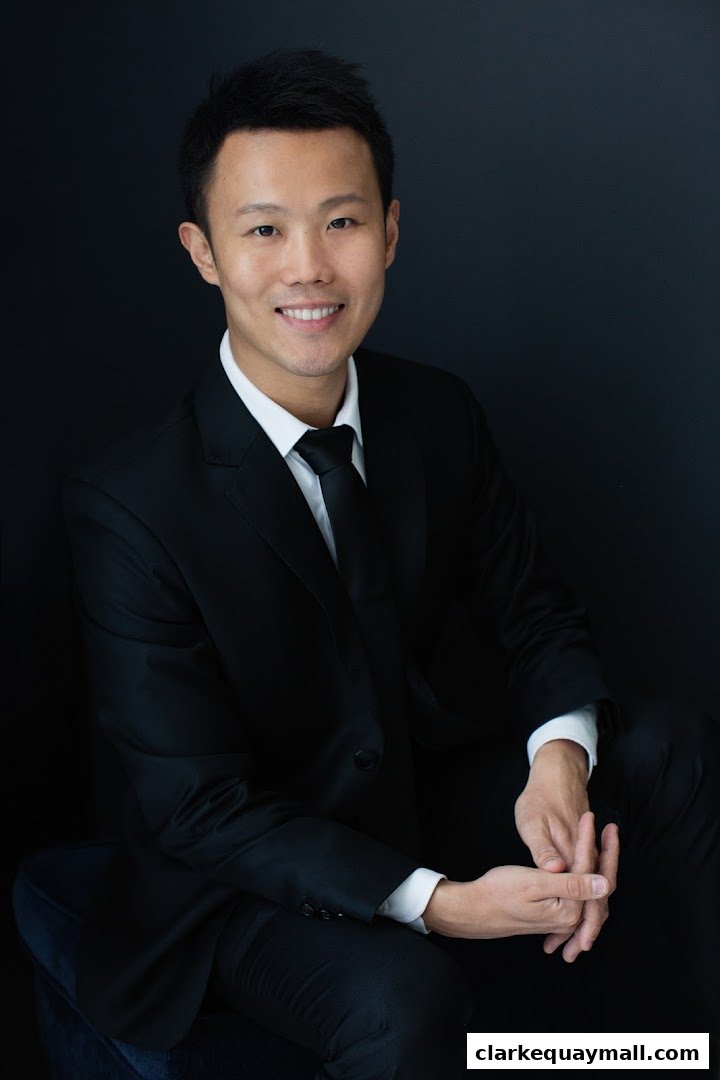 A professional portrait of a man in a black suit and tie against a dark background.