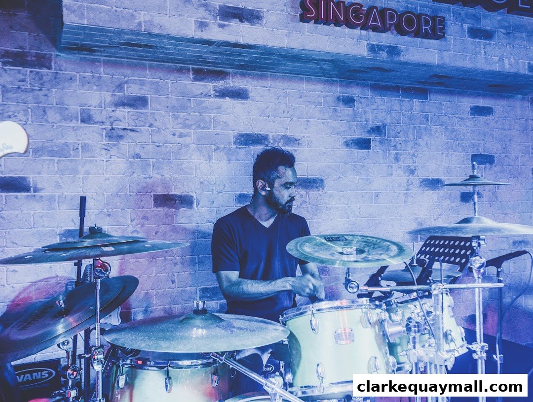 A man plays a drum set in front of a brick wall with a neon 'SINGAPORE' sign.