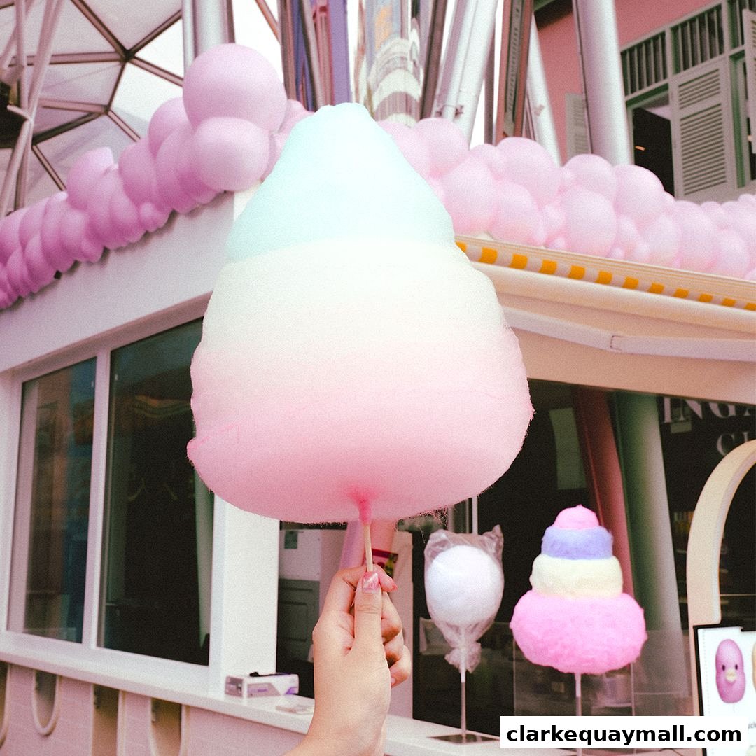 A hand holds a large, colorful cotton candy cone in front of a shop with pink decorations.