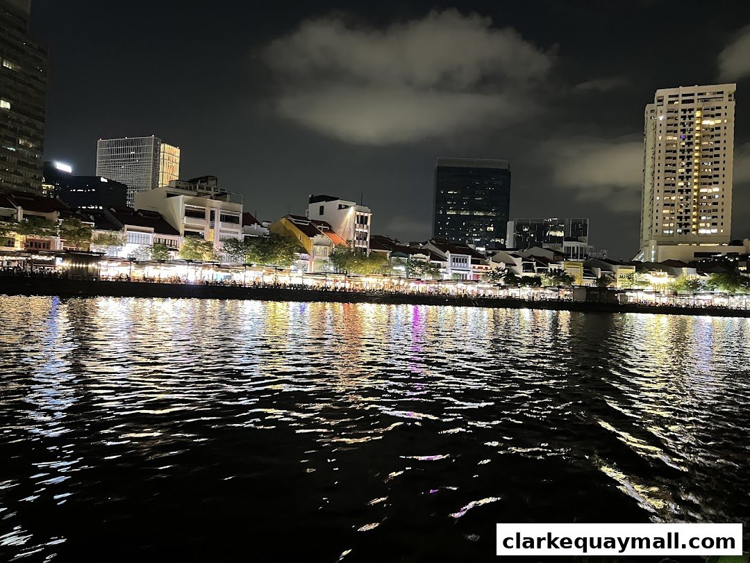 A nighttime view of a bustling riverside promenade with illuminated buildings and reflections on the water.