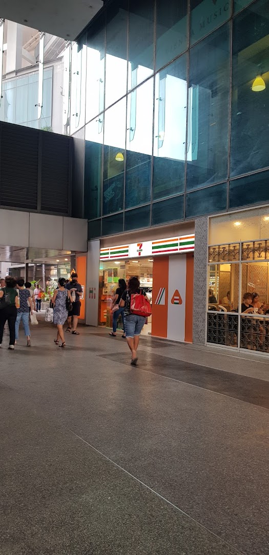 The exterior of a 7-Eleven convenience store with people walking past its entrance in an urban setting.