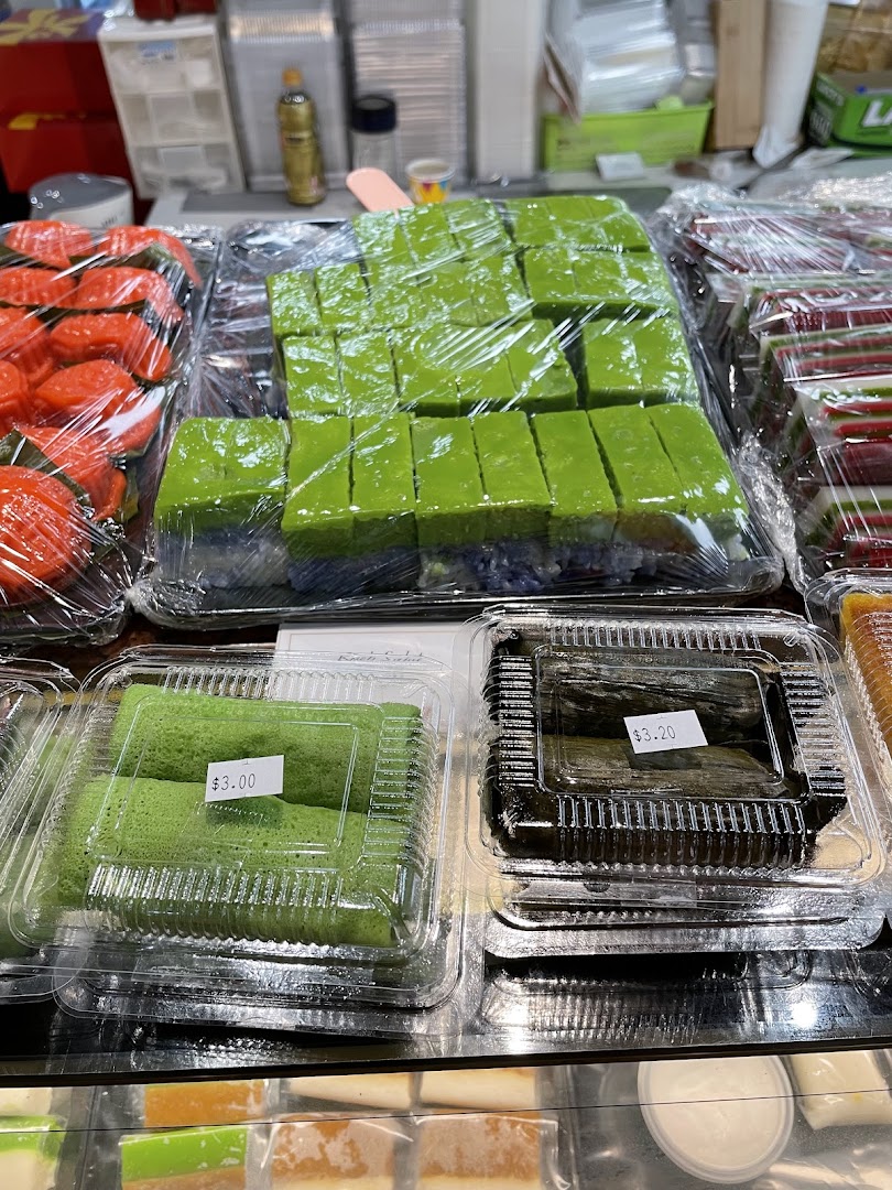 Assorted traditional Indonesian kueh and snacks, including green pandan cakes, displayed in clear packaging.
