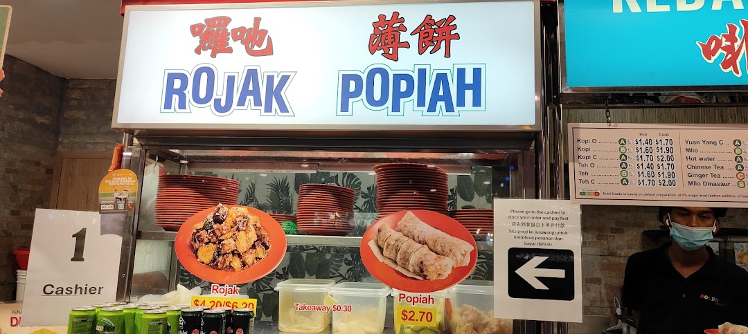 A food stall counter featuring signage for Rojak and Popiah, with a drinks menu and a staff member.