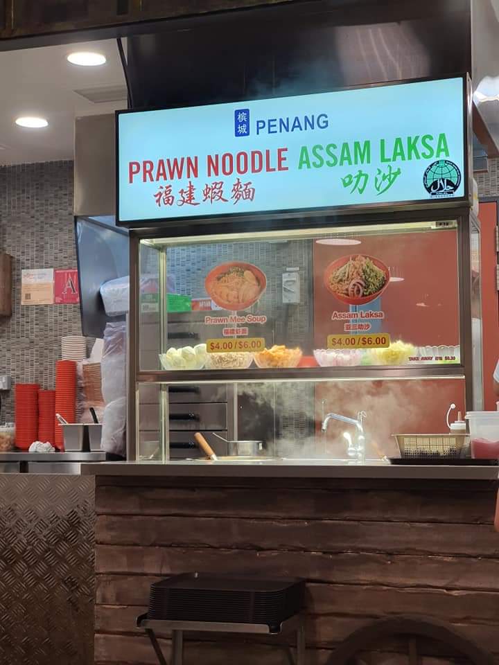 A food stall display board featuring Penang Prawn Noodle and Assam Laksa with prices.
