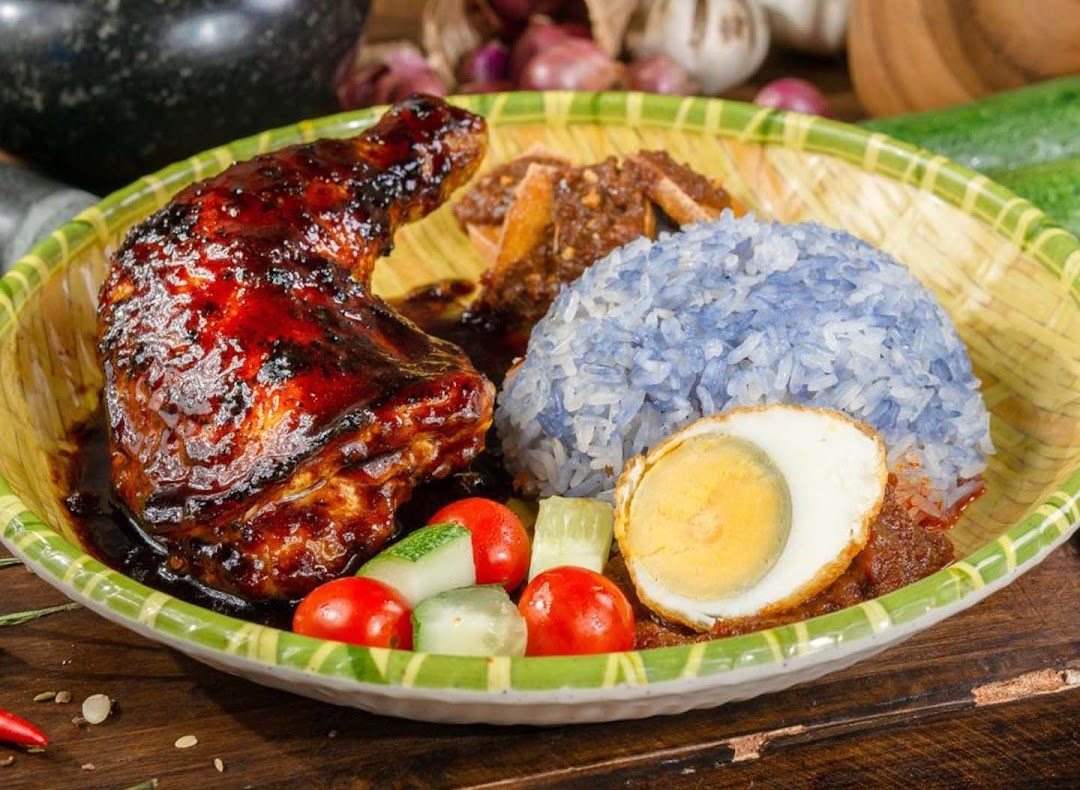 A vibrant plate of Nasi Lemak featuring a glazed chicken leg, blue pea rice, and a fried egg.