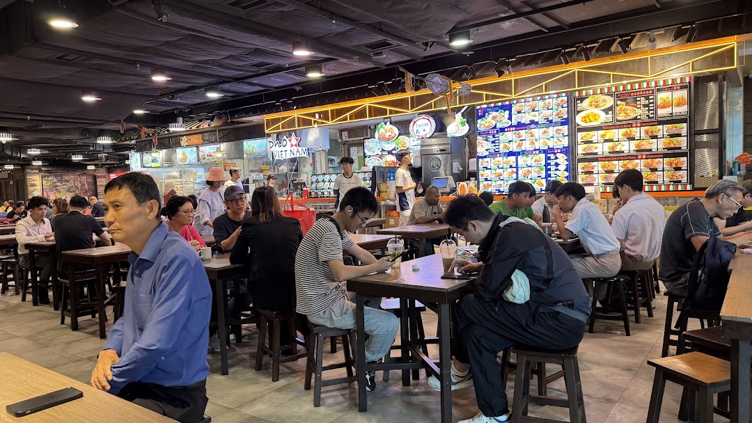 Diners enjoy meals at a bustling food court with various food stalls in the background.