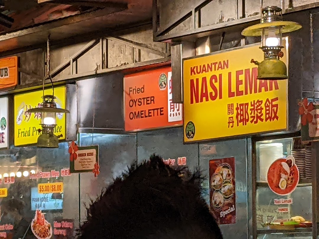 Close-up of a food stall interior featuring signs for Kuantan Nasi Lemak and Fried Oyster Omelette, with vintage lanterns.