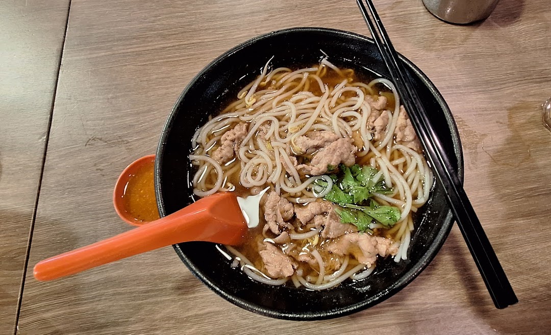 A close-up of a bowl of noodles with sliced meat, bean sprouts, and cilantro, served with a side of chili sauce.