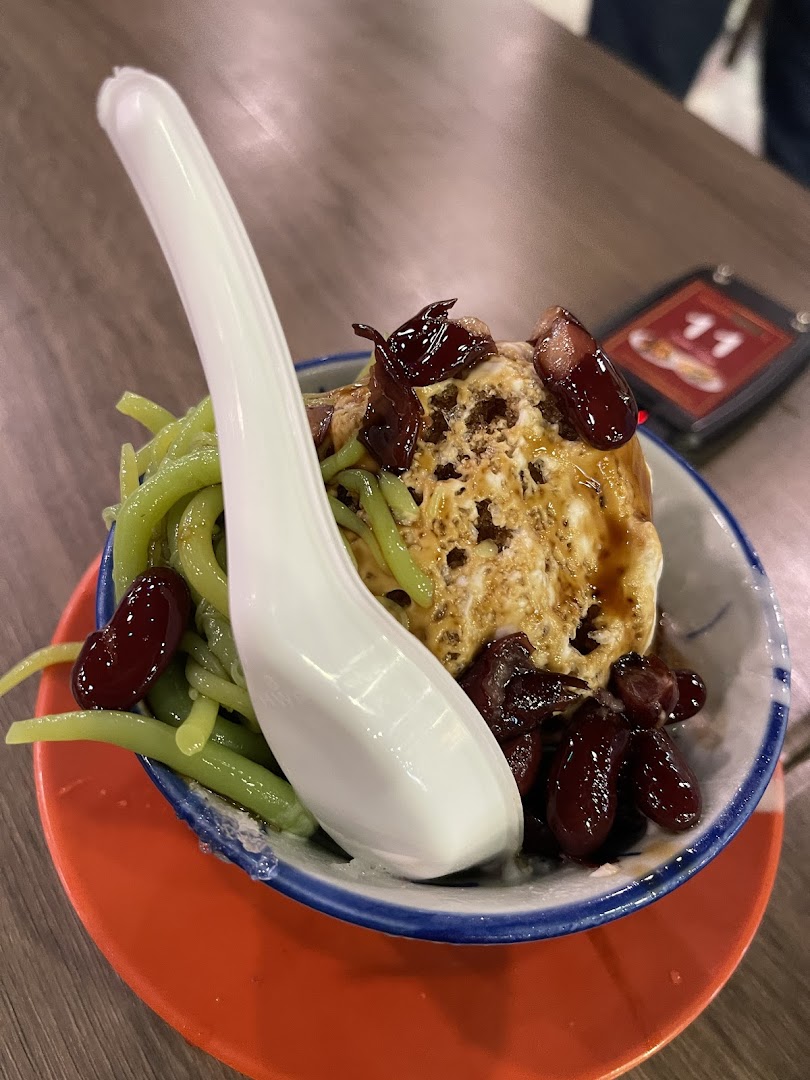 A close-up of a refreshing Cendol dessert with green noodles, red beans, and a white spoon.