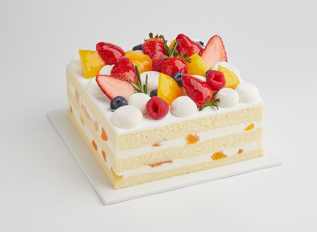 A vibrant square cake topped with fresh berries, mango, and cream on a white background.