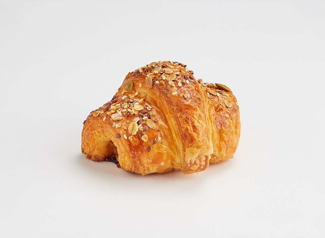 A golden-brown, flaky croissant generously topped with various seeds, presented on a clean white background.